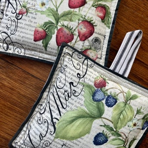May include: Two fabric pot holders with black trim and a white background. The top pot holder features a strawberry design with green leaves and a vintage text pattern. The bottom pot holder features a blackberry design with green leaves and a vintage text pattern. Both pot holders have a black and white striped fabric loop.