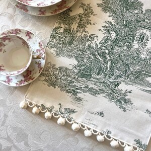 May include: A white table runner with a green toile pattern featuring a pastoral scene with trees, a house, and figures. The runner has a white pom-pom trim.