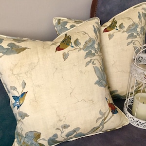 May include: Two cream-colored throw pillows with a floral pattern and colorful birds. The pillows are on a dark blue couch with a white birdcage and a candle.