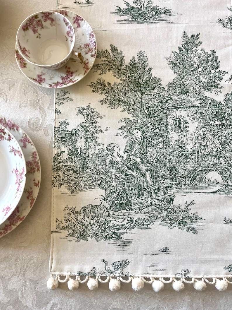 May include: A white table runner with a green toile pattern featuring a pastoral scene with a couple, a bridge, and trees. The runner has a white pom-pom trim.