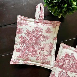 May include: Square, off-white and red Toile de Jouy pot holders with a loop for hanging. The fabric features a pastoral scene with figures, trees, and buildings. The pot holders are approximately 7 inches square.