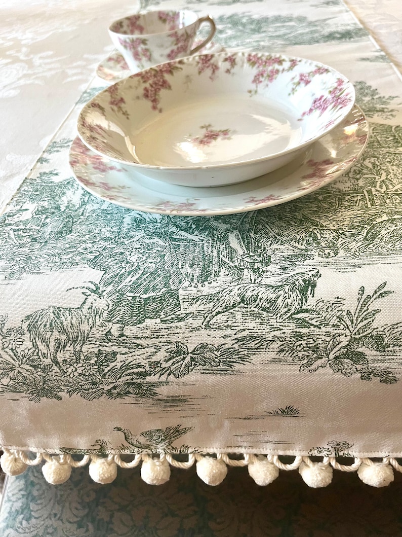 May include: A white table runner with a green and white toile pattern featuring a pastoral scene. The runner has a white pom-pom trim.