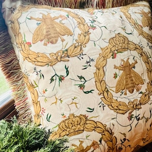 May include: A white pillow with a gold and green floral pattern featuring bees and crowns. The pillow has a brown fringe border.