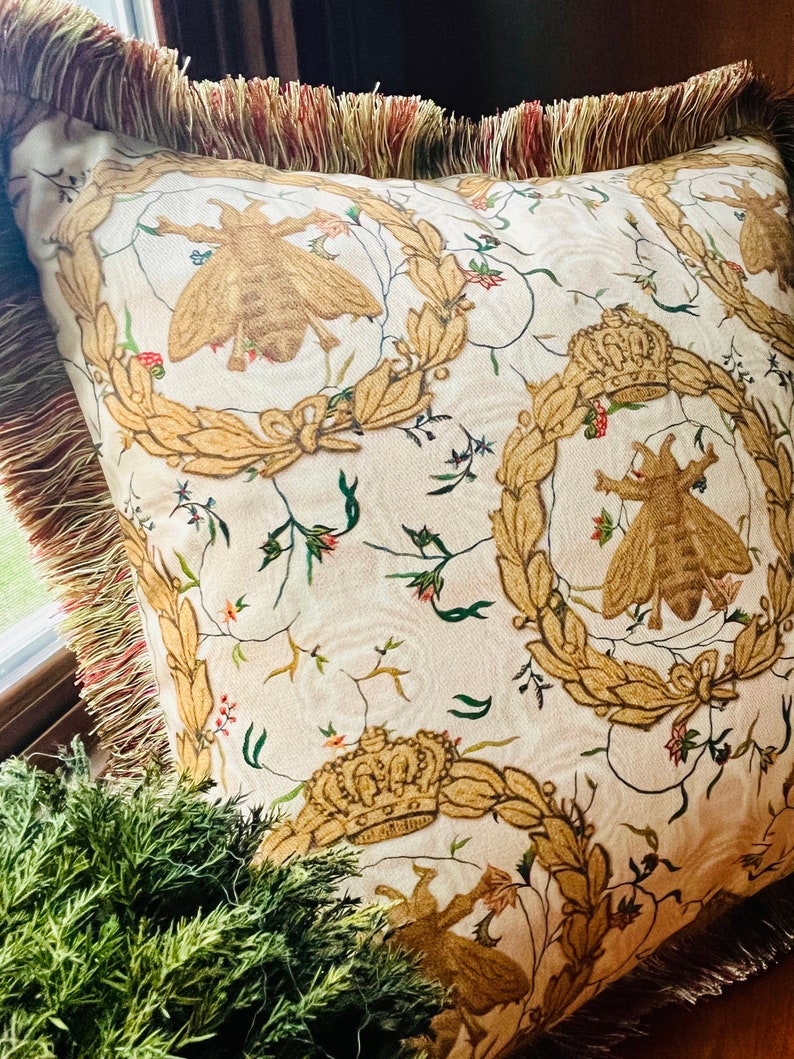 May include: A white pillow with a gold and green floral pattern featuring bees and crowns. The pillow has a brown fringe border.