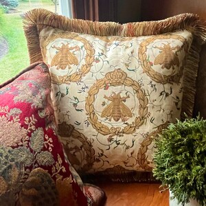 May include: A cream-colored throw pillow with a fringe border features a repeating pattern of bees and floral wreaths. The pillow is on a wooden surface with a red and gold patterned pillow and a small potted plant.