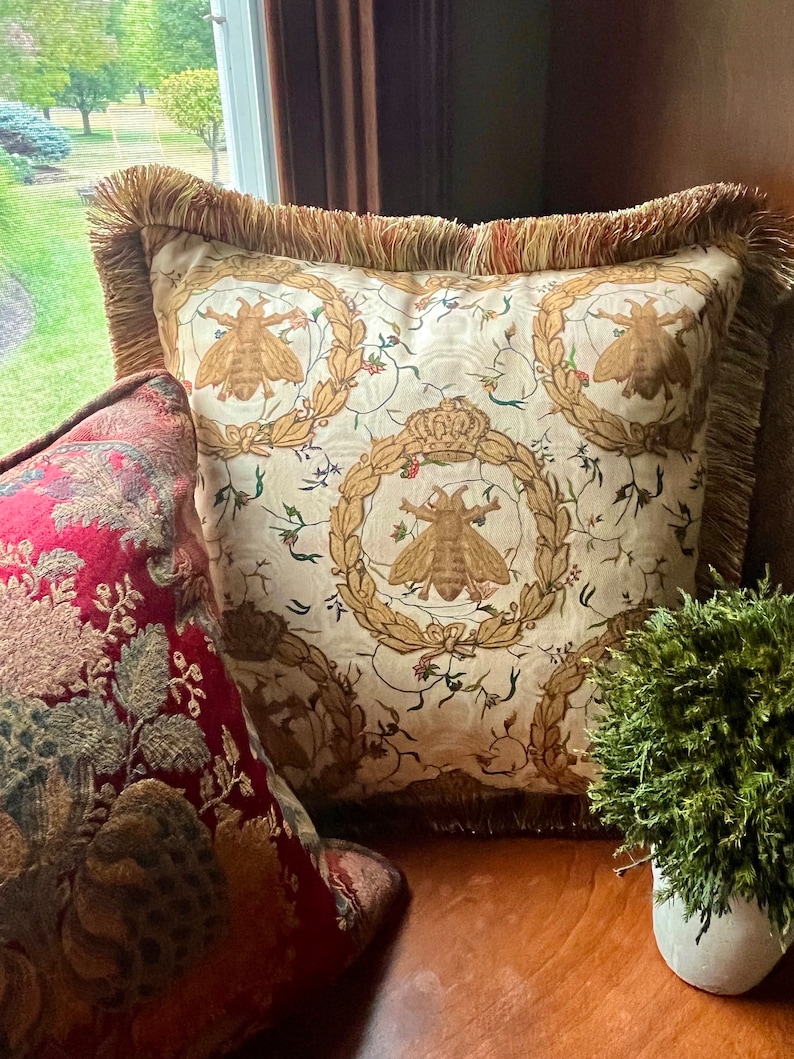 May include: A cream-colored throw pillow with a fringe border features a repeating pattern of bees and floral wreaths. The pillow is on a wooden surface with a red and gold patterned pillow and a small potted plant.