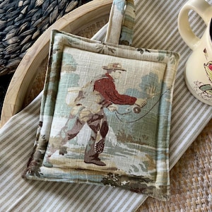 May include: Square fabric pot holder with a handle. The center features a vintage illustration of a person fly fishing in a river, with a red jacket and hat. The border and handle have a coordinating floral pattern.