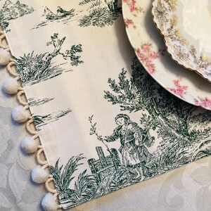 May include: A white table runner with a green toile pattern featuring a pastoral scene. The runner has a fringe of white pom-poms along one edge.