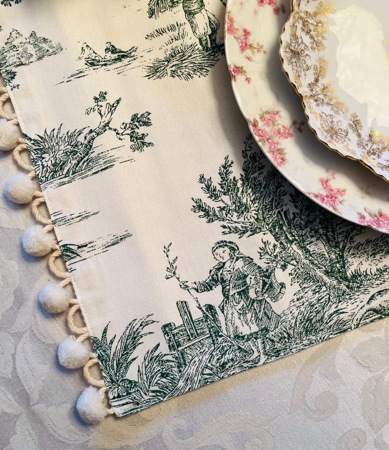 May include: A white table runner with a green toile pattern featuring a pastoral scene. The runner has a fringe of white pom-poms along one edge.