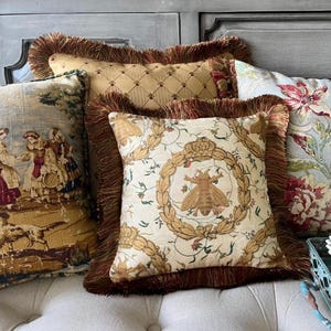 May include: Three decorative throw pillows with fringed edges. The pillows are arranged on a light beige couch. The pillow in the front features a white background with a gold bee and floral design. The middle pillow has a gold background with a diamond pattern. The pillow in the back has a blue and white floral pattern.