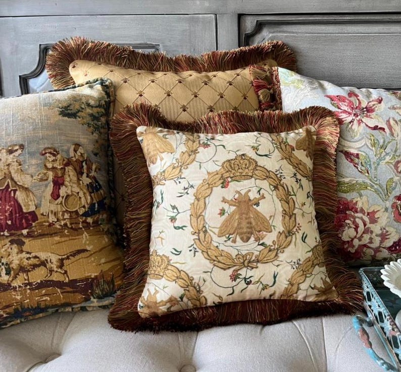 May include: Three decorative throw pillows with fringed edges. The pillows are arranged on a light beige couch. The pillow in the front features a white background with a gold bee and floral design. The middle pillow has a gold background with a diamond pattern. The pillow in the back has a blue and white floral pattern.