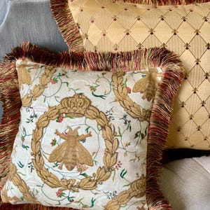 May include: Two decorative throw pillows with fringed edges. The pillow in the foreground features a white background with a repeating pattern of a bee, a crown, and floral elements. The pillow in the background is a solid gold color with a diamond pattern.