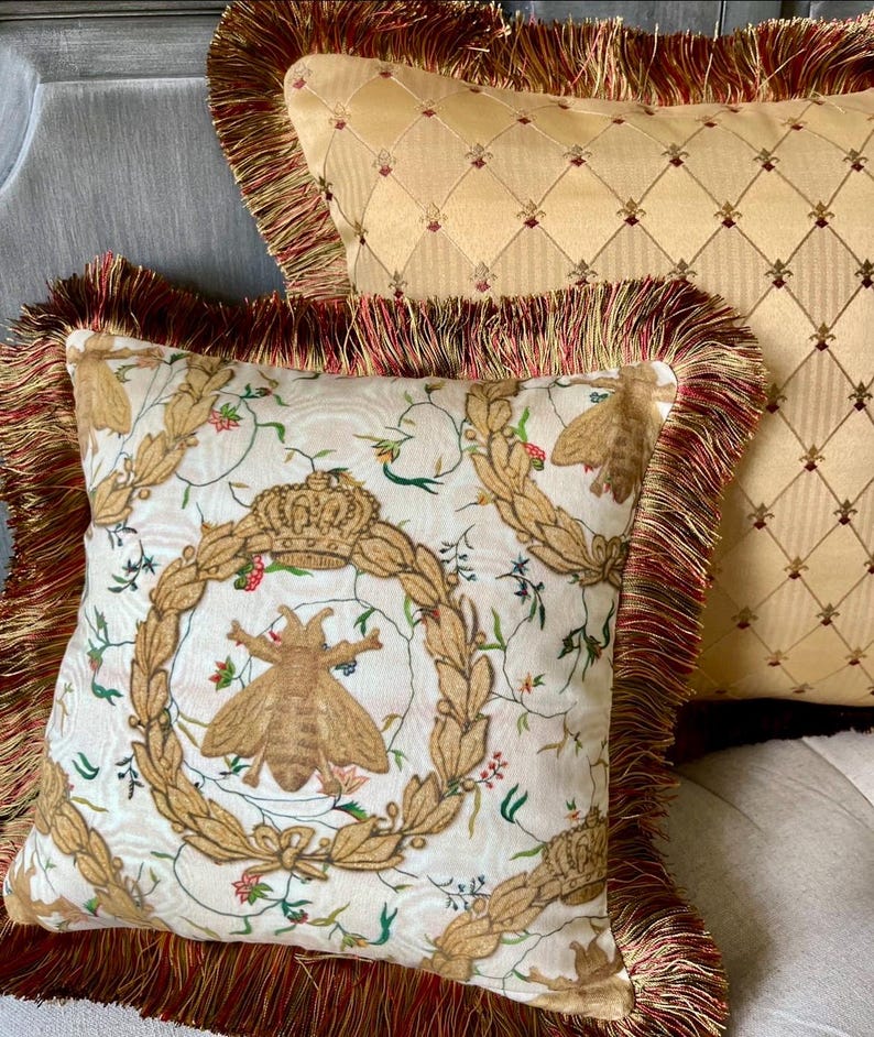 May include: Two decorative throw pillows with fringed edges. The pillow in the foreground features a white background with a repeating pattern of a bee, a crown, and floral elements. The pillow in the background is a solid gold color with a diamond pattern.