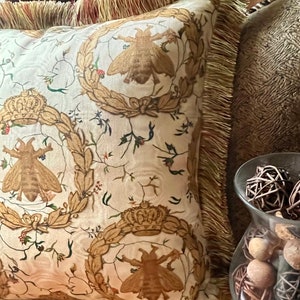 May include: A decorative throw pillow with a white background and a repeating pattern of gold crowns, bees, and floral designs. The pillow has a gold fringe border.