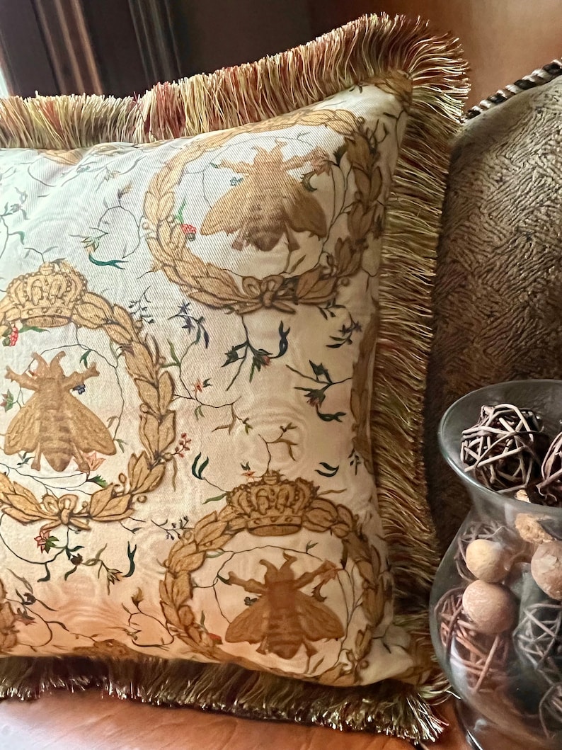 May include: A decorative throw pillow with a white background and a repeating pattern of gold crowns, bees, and floral designs. The pillow has a gold fringe border.