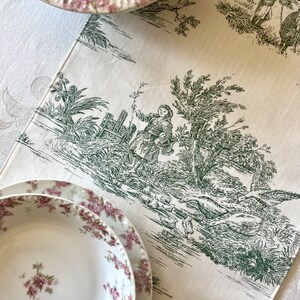 May include: A white table runner with a green toile pattern featuring a pastoral scene with a person and birds. The runner is decorated with a green and white floral pattern.