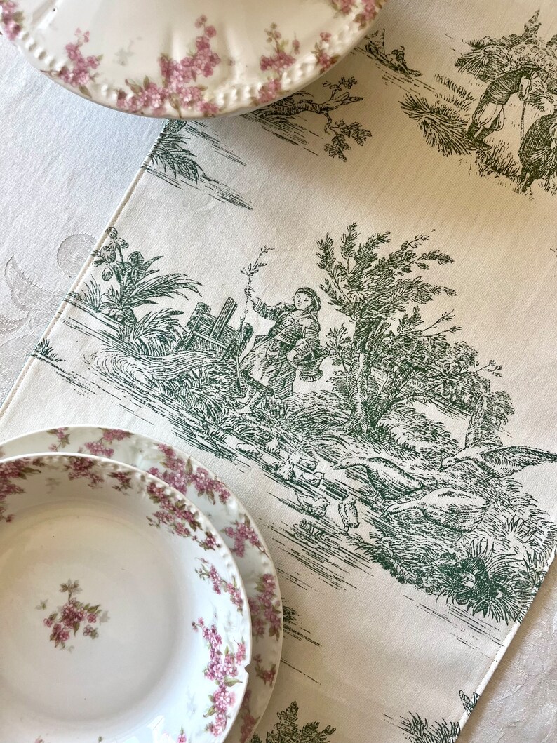 May include: A white table runner with a green toile pattern featuring a pastoral scene with a person and birds. The runner is decorated with a green and white floral pattern.