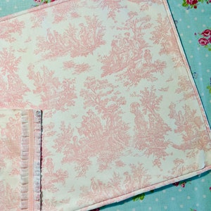 A white pillow cover with a pink toile pattern. The pattern features a repeating design of trees, flowers, and people in a pastoral setting. The pillow cover has a corded welt on edge and is sewn with a light pink thread.