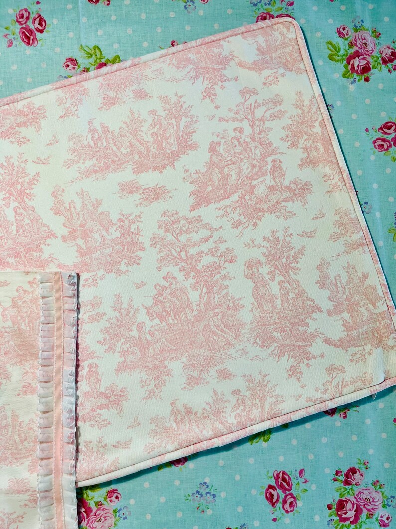 A white pillow cover with a pink toile pattern. The pattern features a repeating design of trees, flowers, and people in a pastoral setting. The pillow cover has a corded welt on edge and is sewn with a light pink thread.