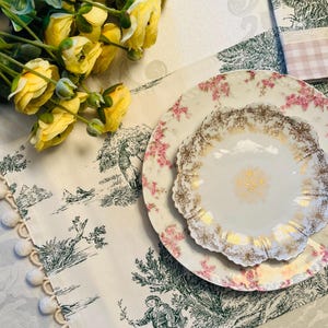 May include: A white porcelain plate with a delicate floral pattern and gold accents. The plate is set on a white tablecloth with a green and white floral design. The tablecloth has a white pom-pom trim.