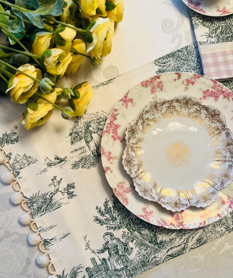 May include: A white porcelain plate with a delicate floral pattern and gold accents. The plate is set on a white tablecloth with a green and white floral design. The tablecloth has a white pom-pom trim.