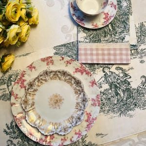 May include: A white porcelain dinner plate with a gold and pink floral design. The plate is set on a white tablecloth with a green and white floral pattern.