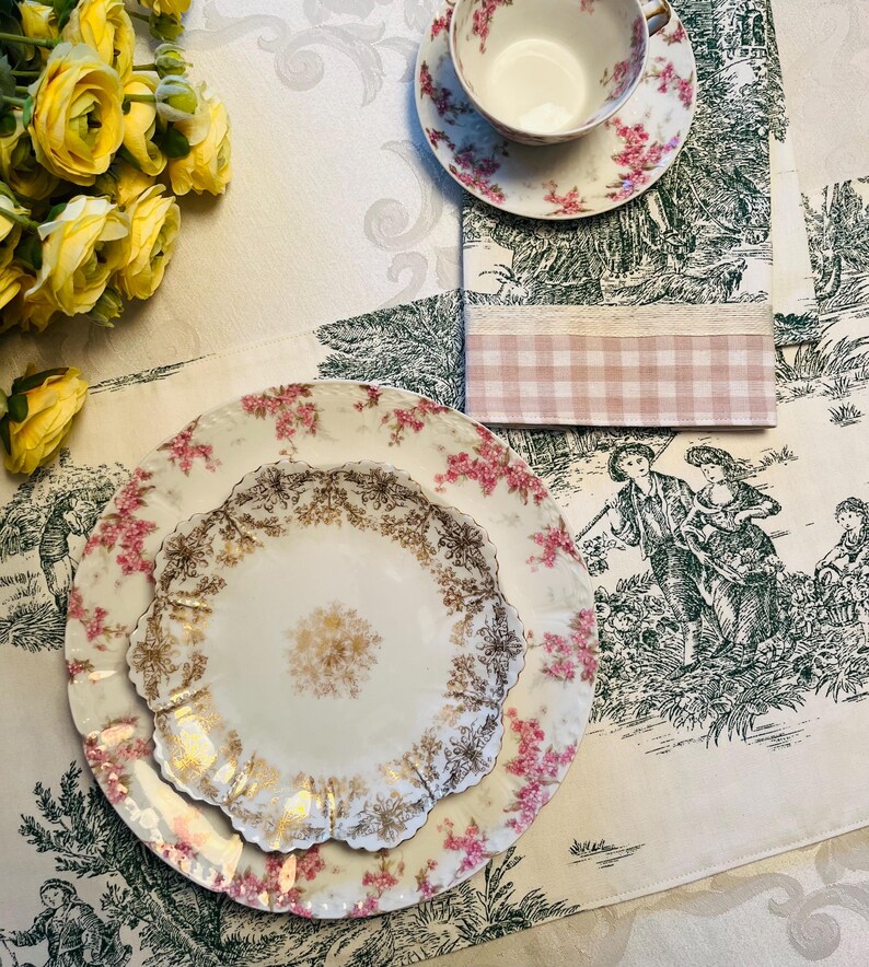 May include: A white porcelain dinner plate with a gold and pink floral design. The plate is set on a white tablecloth with a green and white floral pattern.