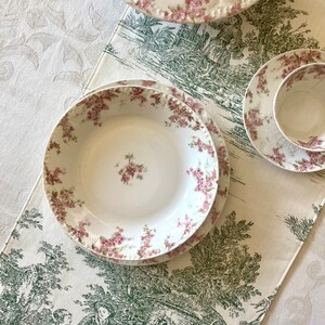 May include: A set of three white porcelain plates with a floral pattern in shades of pink and green. The plates are on a white tablecloth with a green floral pattern.