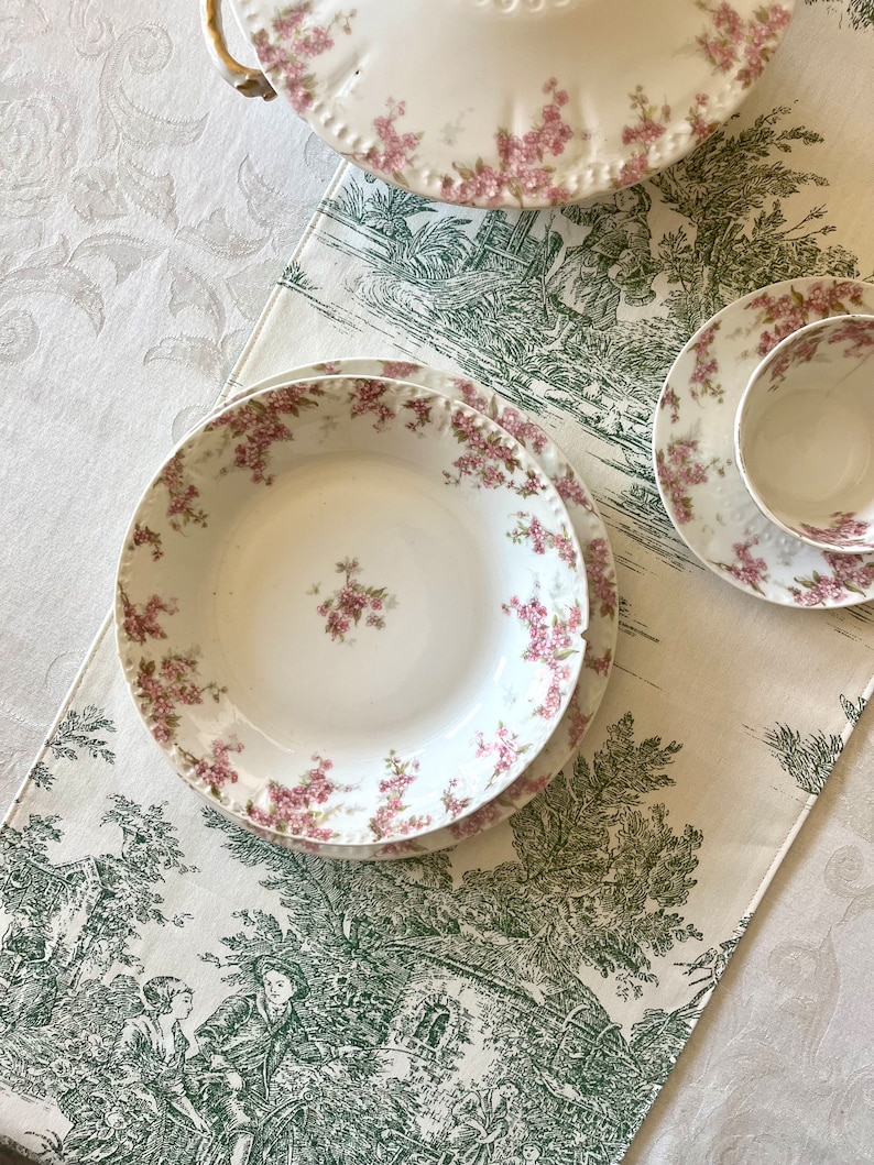 May include: A set of three white porcelain plates with a floral pattern in shades of pink and green. The plates are on a white tablecloth with a green floral pattern.