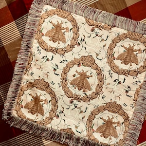 May include: A square throw pillow with a cream-colored fabric featuring a repeating pattern of bees, crowns, and floral motifs. The pillow has a brown and cream fringe border.