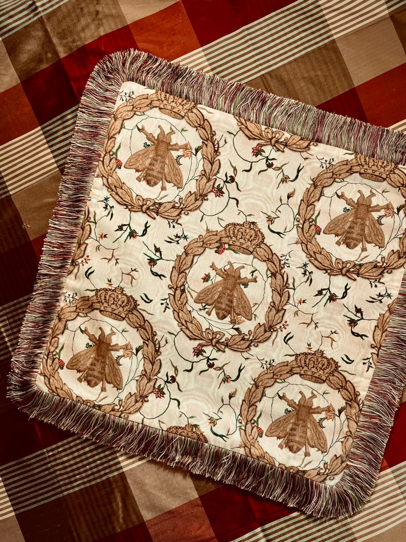 May include: A square throw pillow with a cream-colored fabric featuring a repeating pattern of bees, crowns, and floral motifs. The pillow has a brown and cream fringe border.