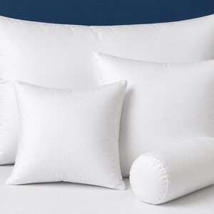 May include: A selection of white pillows in different shapes and sizes. The set includes a rectangular pillow, two square pillows, and a cylindrical bolster. The pillows are set against a dark blue backdrop.