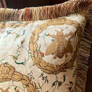 May include: A cream-colored pillow with a gold and brown floral pattern. The pillow has a gold fringe trim.