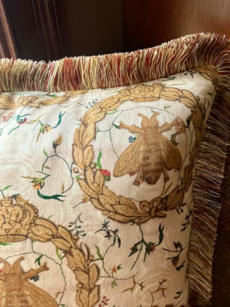 May include: A cream-colored pillow with a gold and brown floral pattern. The pillow has a gold fringe trim.