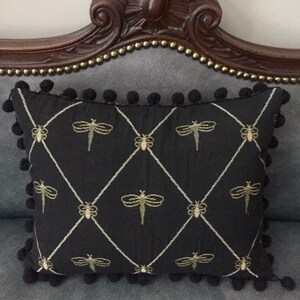 May include: A black rectangular cushion featuring gold dragonfly embroidery in a diamond pattern. The cushion is trimmed with black pom-poms and rests on a grey upholstered surface.