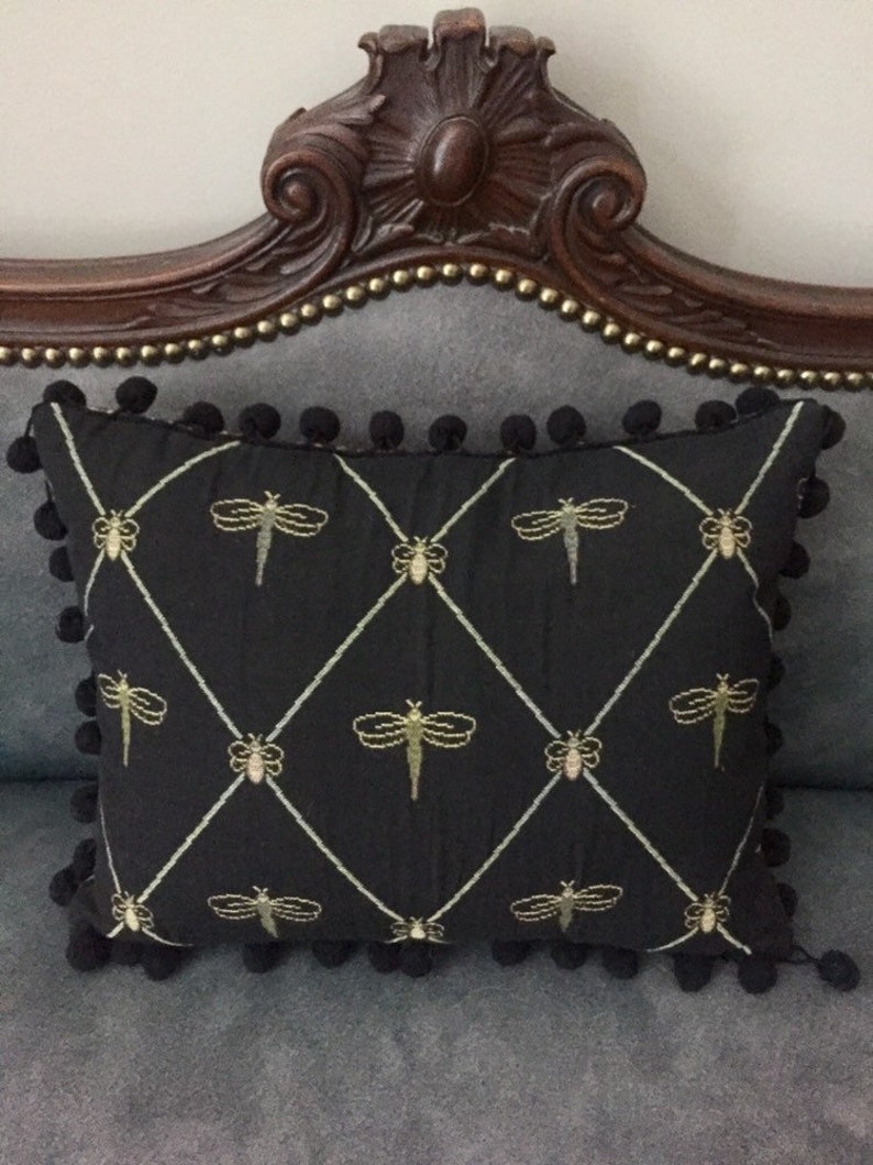 May include: A black rectangular cushion featuring gold dragonfly embroidery in a diamond pattern. The cushion is trimmed with black pom-poms and rests on a grey upholstered surface.