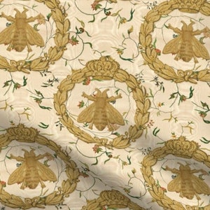 May include: A white fabric with a repeating pattern of gold bees, crowns, and floral vines. The bees are stylized and have a golden color.