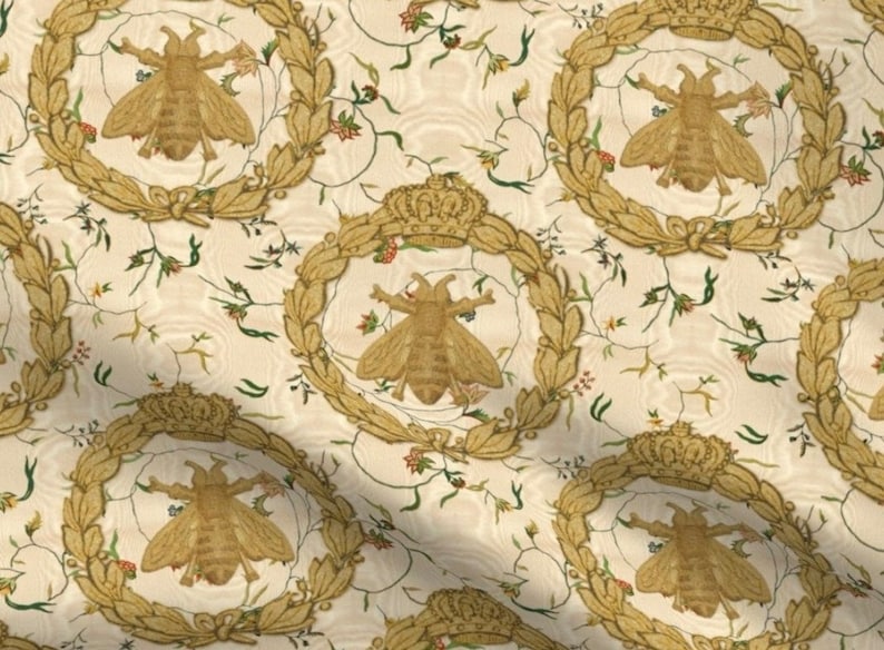 May include: A white fabric with a repeating pattern of gold bees, crowns, and floral vines. The bees are stylized and have a golden color.