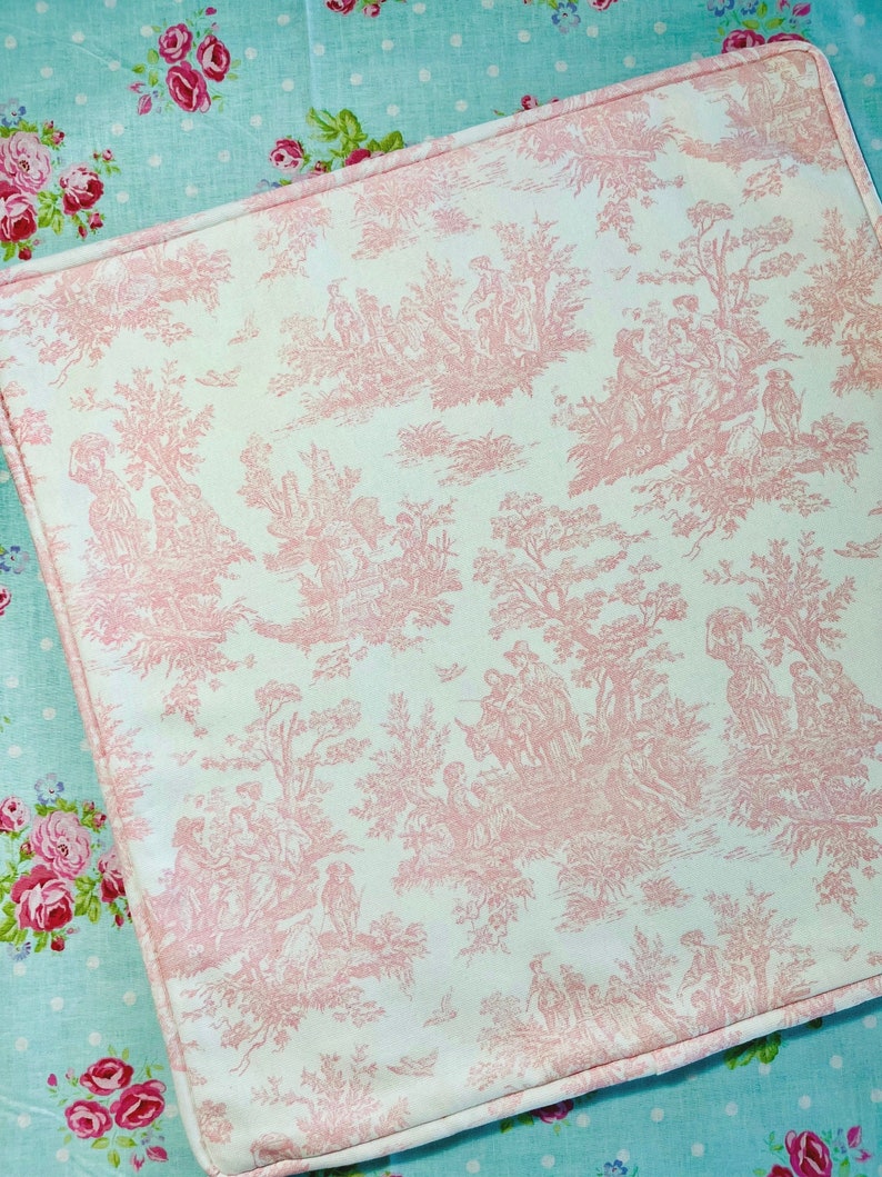A white pillow cover with a pink toile pattern. The pattern features a repeating design of trees, flowers, and people in a pastoral setting. The pillow cover has a corded welt on edge and is sewn with a light pink thread.