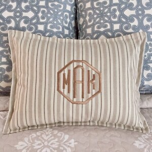 May include: A rectangular cushion with vertical beige and brown stripes. The centre has an embroidered monogram within a geometric shape. Two blue and white patterned cushions are in the background. The cushion is on a bed with a floral pattern.