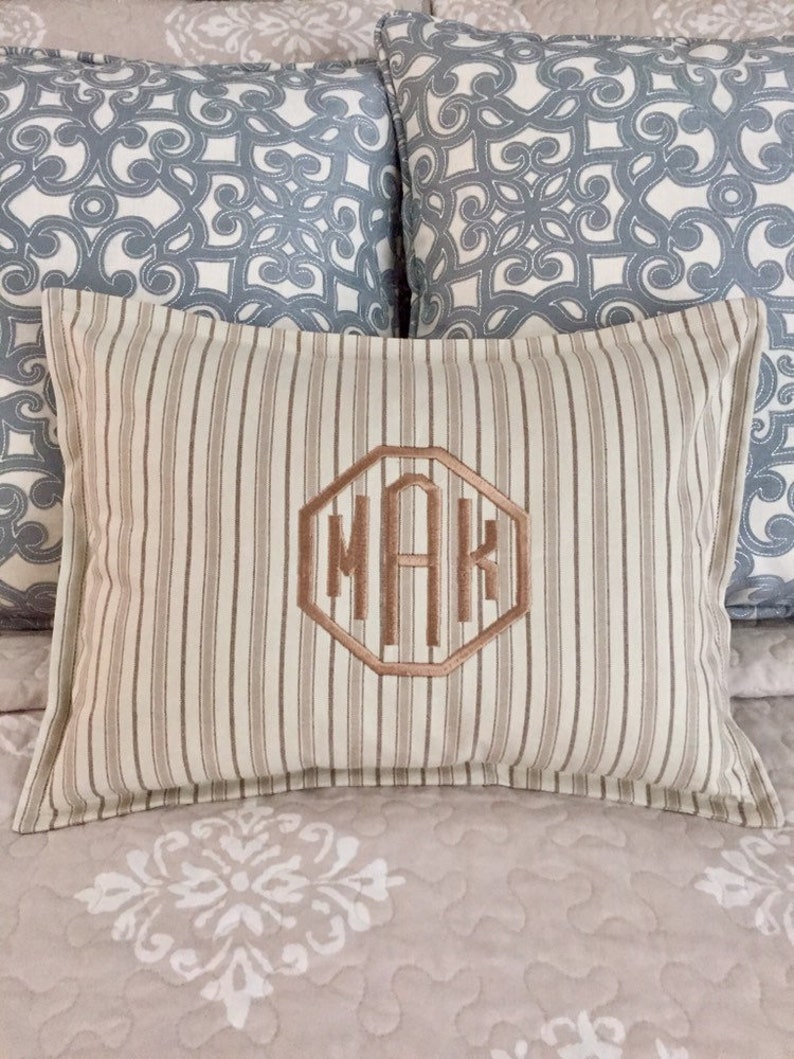 May include: A rectangular cushion with vertical beige and brown stripes. The centre has an embroidered monogram within a geometric shape. Two blue and white patterned cushions are in the background. The cushion is on a bed with a floral pattern.
