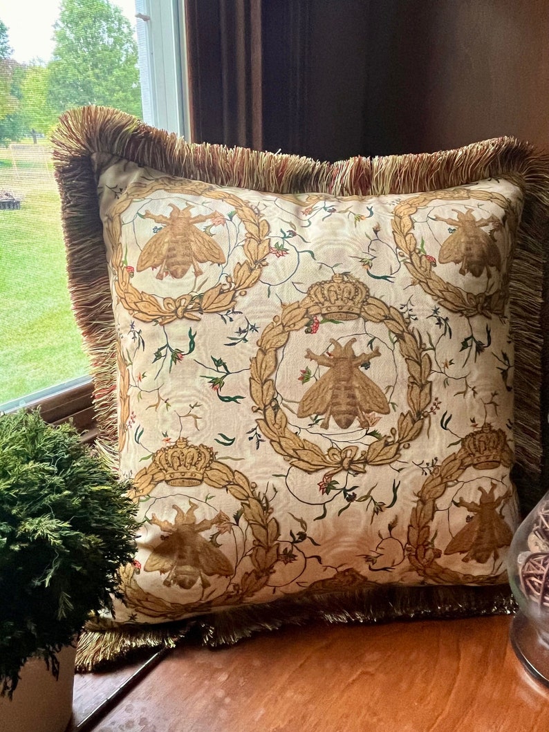 May include: A cream-colored throw pillow with a gold fringe border features a repeating pattern of bees and crowns. The bees are depicted in a vintage style with intricate details. The pillow is perfect for adding a touch of elegance to any room.