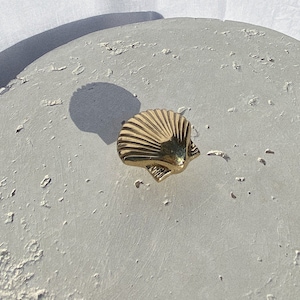 May include: A gold-coloured seashell-shaped decorative object. The object has a textured surface with detailed ridges, and is set against a light grey, textured background. The seashell is a decorative element.