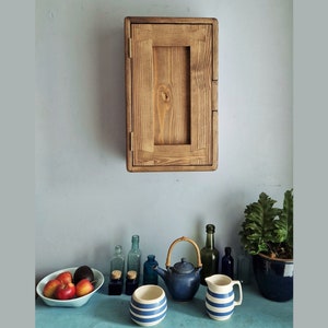 May include: A wooden wall-mounted cabinet with a single door. The cabinet is made of light brown wood and has a simple design. The cabinet is mounted on a light blue wall. There are several bottles, a teapot, and a pitcher on a blue countertop in front of the cabinet.