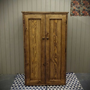 May include: A tall, rectangular wooden cabinet with a medium-brown stained finish and visible wood grain. The cabinet features two vertical doors, each equipped with a small, round wooden knob centred on the door panel.