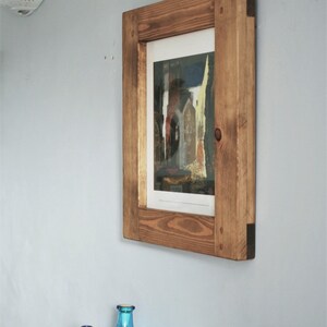 May include: A framed print of a building with a wooden frame. The frame is made of light brown wood and is mounted on a light blue wall. The print features a colorful image of a building.