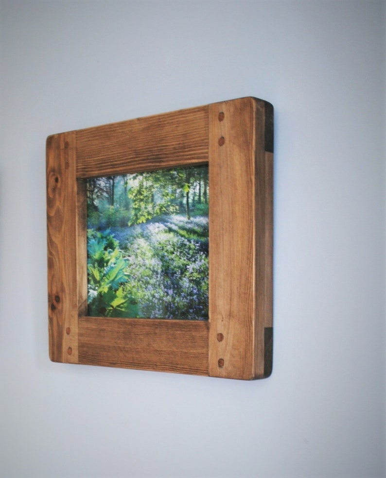 May include: Square wooden picture frame with a natural wood finish. The frame surrounds a landscape image of a forest with green foliage and sunlight. The frame is mounted on a light blue wall.