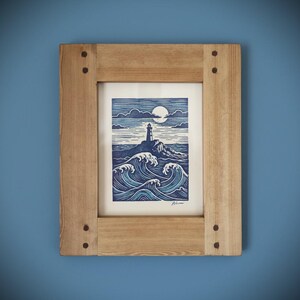 May include: A framed print depicting a lighthouse on a rocky outcrop, with waves and a moonlit sky. The artwork uses blue and white tones, encased in a natural wood frame with visible fixings. The image evokes a coastal theme.