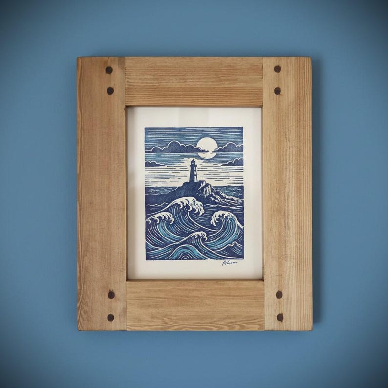 May include: A framed print depicting a lighthouse on a rocky outcrop, with waves and a moonlit sky. The artwork uses blue and white tones, encased in a natural wood frame with visible fixings. The image evokes a coastal theme.