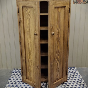 May include: A tall, rectangular wooden cupboard with a medium brown stained finish and visible wood grain. It features two hinged doors with small round knobs, opening to reveal five internal shelves stacked vertically inside the unit.